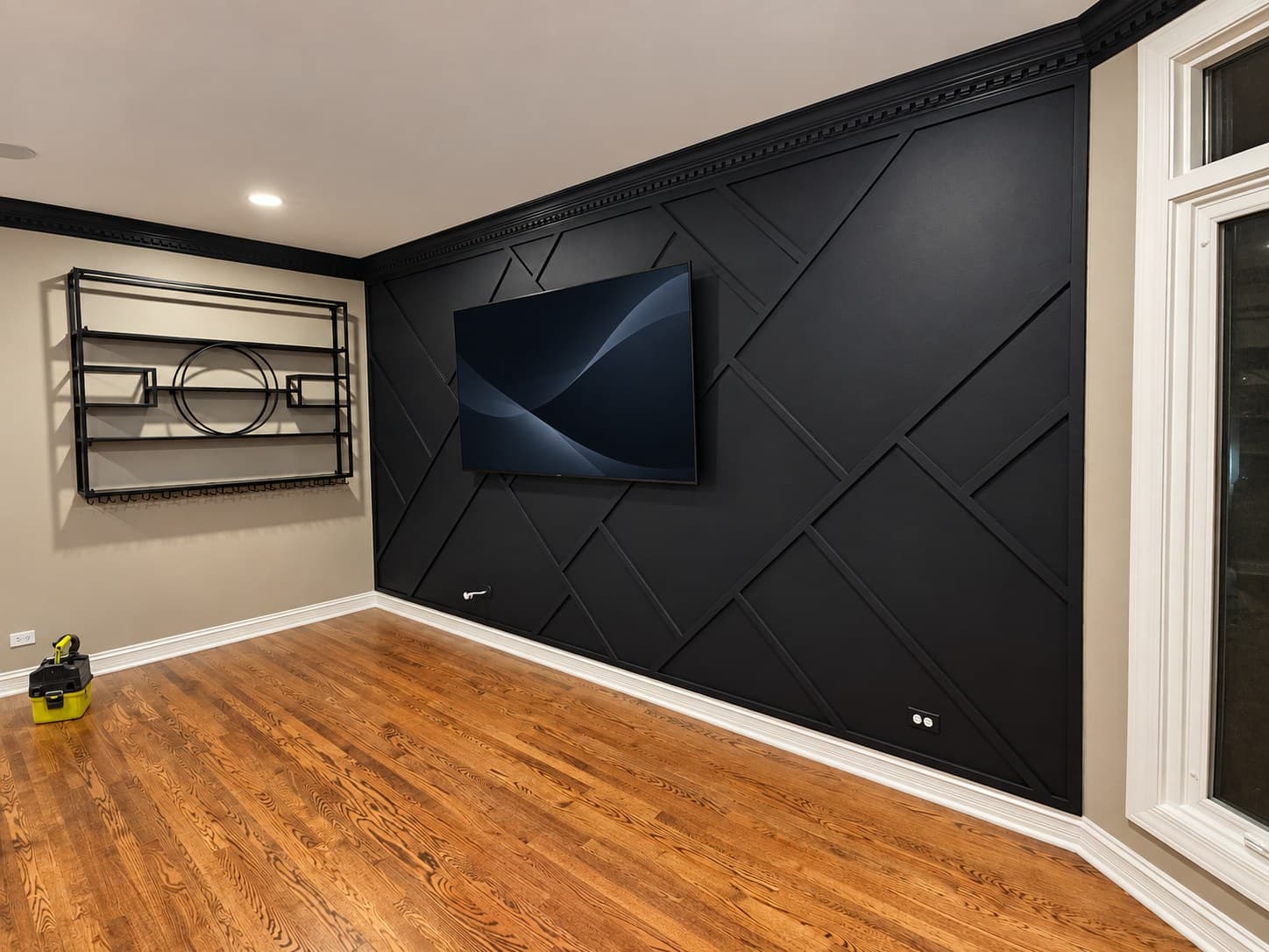 Black geometric trim accent wall behind a mounted TV, with black crown molding and a wall-mounted metal shelf sculpture