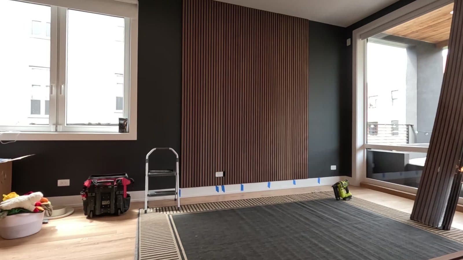 Walnut wood slat acoustic panel on a dark living room wall between two large windows, during installation